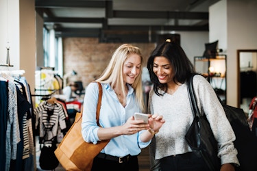  Two women stand in a clothing shop and look down at a smartphone. The woman on the left has long blonde hair and is wearing a light blue button-up shirt. She has a tan handbag over one shoulder. She is holding the smartphone and pointing at something on its screen. The woman on the right has long, dark hair and is wearing a pale gray sweater. She has a black handbag over one shoulder and she looks down at the smartphone with a smile. 